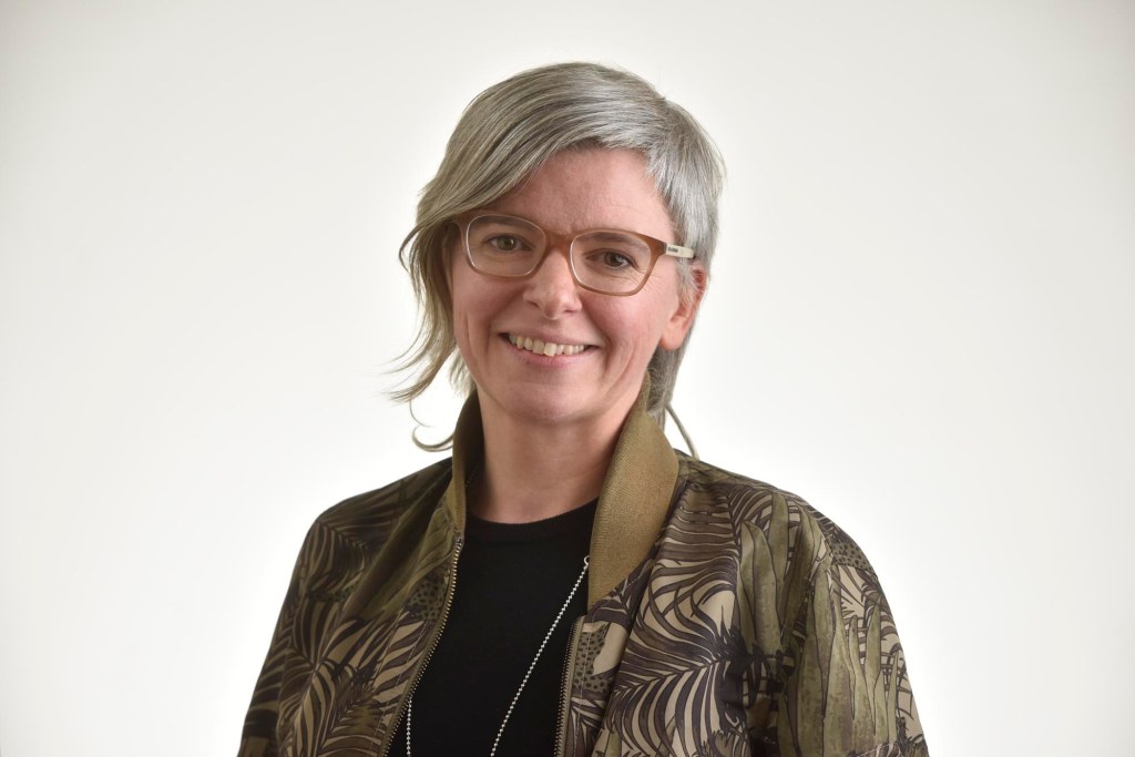 A photograph of Alys, a white woman with glasses and grey hair, smiling at the camera. She is wearing a jacket patterned with green leaves.