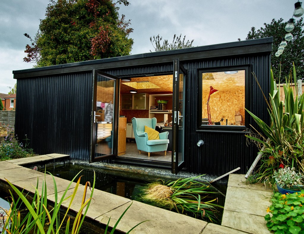 A photograph of the black studio in my garden, behind a pond with a bridge across. The warm lights are glowing and there's a comfy turquoise armchair in the open window.