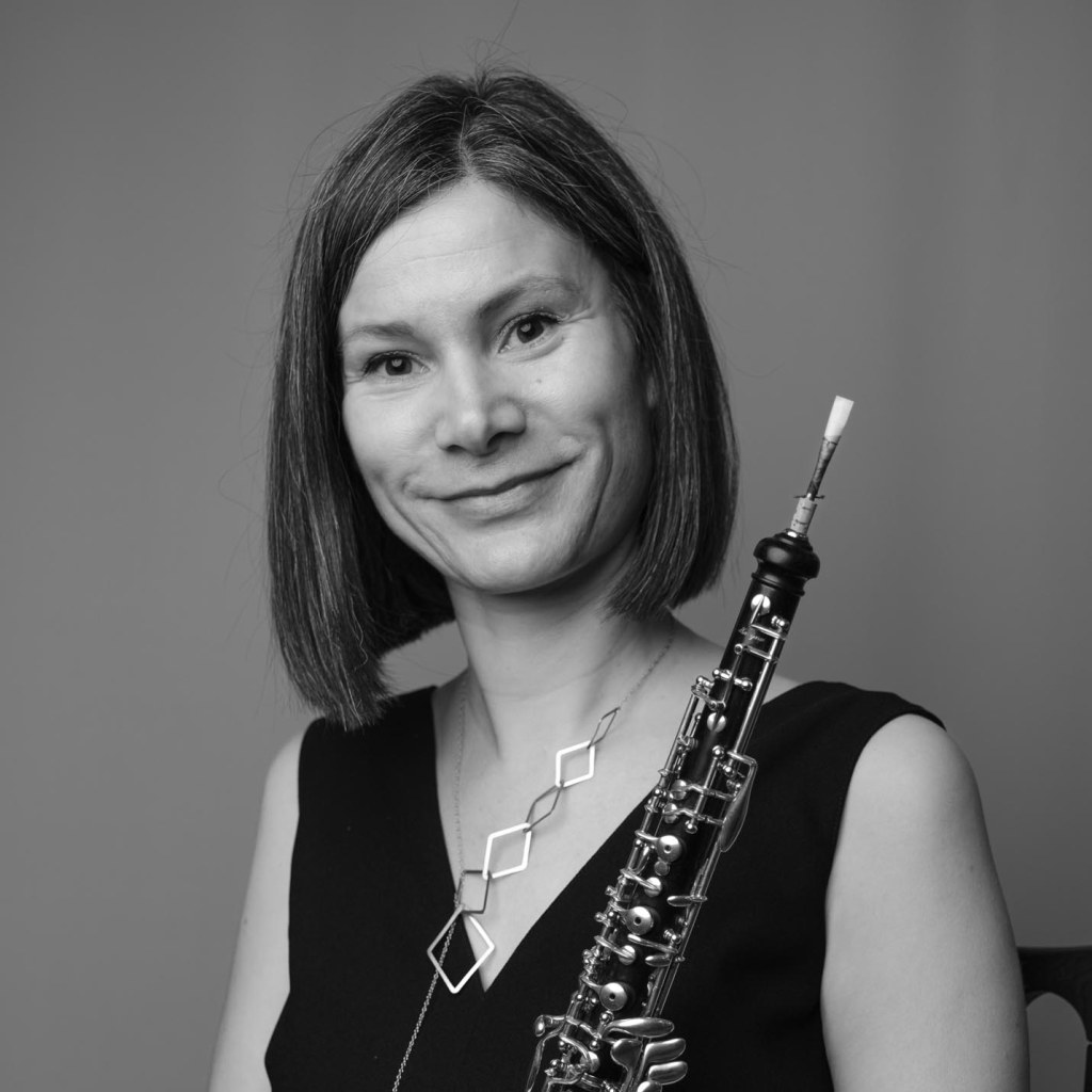 A black and white image of a white woman with dark hair, looking at the camera and holding an oboe.