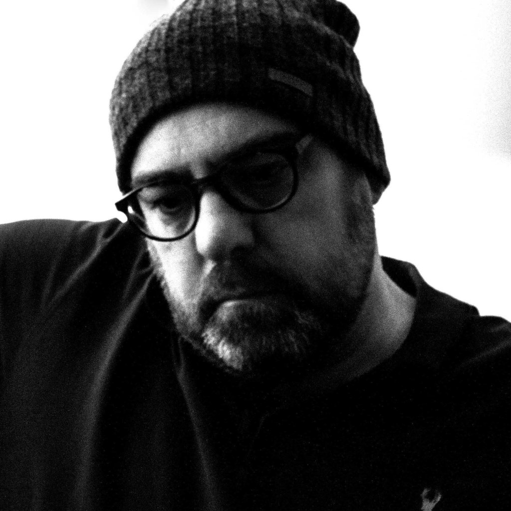 A black and white image of a white man with a beard, wearing glasses and a woolly hat. He is looking serious.