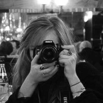A black and white image of a white woman with long hair, with glowing lights of a pub interior behind her. Her face is obscured by the camera she is looking through.