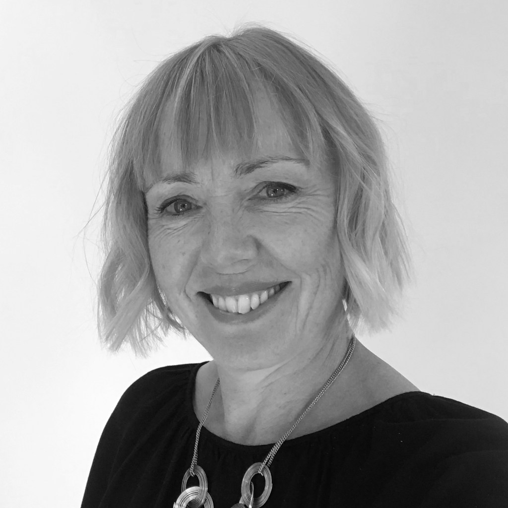 A black and white image of a white woman with blonde hair, smiling. She is wearing a silver necklace with interlocking rings.