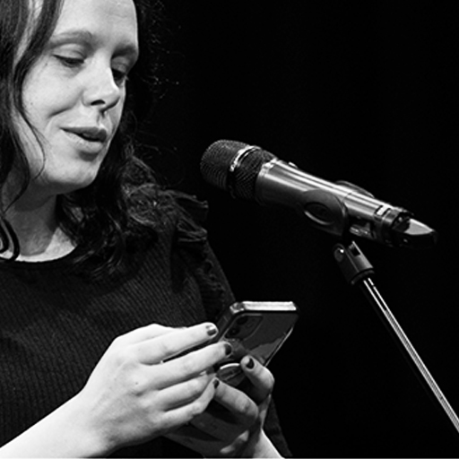 A black and white photograph of a white woman with dark wavy hair, in front of a microphone. She is looking down at a phone and reading poetry. 